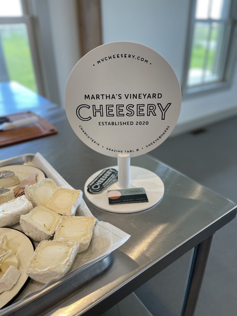 The Martha's Vineyard Cheesery sign, stickers, and business cards next to a tray of bloomy rind cheese.